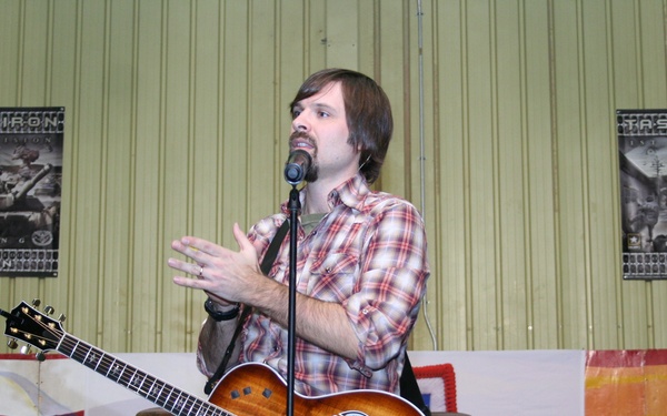 A male musician is performing on stage at what appears to be a military event, singing into a microphone while holding an acoustic guitar. Behind him are two identical 'Task Force Iron - 1st Armored Division' banners, along with a patriotic mural featuring an American flag and military helicopters. A second musician is partially visible on the right side of the frame near a drum kit.