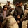 Iraqi Security Force take lead during Ashura movement in Kadhimiyah The image shows a security checkpoint scene where uniformed soldiers in desert camouflage are conducting searches or checks on a group of civilians, including an elderly man, a young boy, and several adult men. One soldier is bent down performing a pat-down or bag check while another stands nearby. The setting appears to be an outdoor area near a chain-link fence with barbed wire.