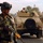 Iraqi Security Force take lead during Ashura movement in Kadhimiyah A soldier in camouflage uniform and black helmet stands in the foreground holding an AK-pattern rifle, appearing to conduct a security patrol or checkpoint operation. Behind him is a tan-colored military Humvee with a second soldier visible in the turret position. The setting appears to be an urban street environment with palm trees and low-rise buildings visible in the background.