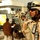 Iraqi Security Force take lead during Ashura movement in Kadhimiyah The image shows armed soldiers in desert camouflage uniforms conducting what appears to be a security or checkpoint operation in an urban Middle Eastern setting. The foreground soldier is equipped with a tactical helmet with mounted goggles, sunglasses, a radio, and handcuffs visible on their vest. Civilians are present in the background near what appears to be a checkpoint area with traffic cones and concertina wire.