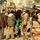 Iraqi Security Force take lead during Ashura movement in Kadhimiyah The image shows a security checkpoint or patrol in an urban Middle Eastern setting, likely Iraq, where armed soldiers in desert camouflage uniforms are monitoring a crowd of civilians. Two soldiers wearing helmets, body armor, and carrying rifles are visible in the foreground, with a large crowd of civilians including women in black abayas in the background. The scene appears to be during a public gathering or religious procession, with barbed wire and traffic cones used as crowd control barriers.