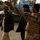 Iraqi Security Force take lead during Ashura movement in Kadhimiyah The image shows several men on a street in what appears to be an urban Middle Eastern setting. One man wearing desert camouflage uniform and a black beanie hat with a tactical vest is gesturing with his arm extended, appearing to direct or signal others. The background shows palm trees, a blue building, rubble, and a parked dark sedan.