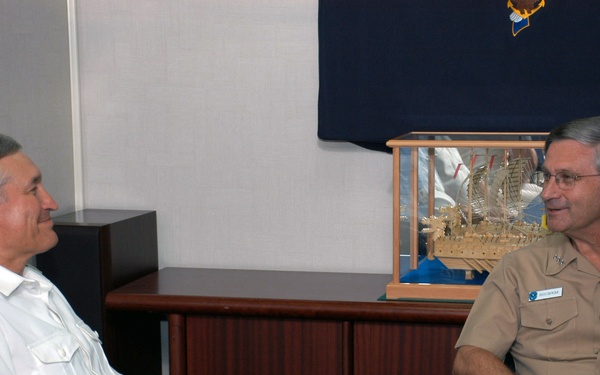 Two military officers are seated at a conference table engaged in conversation. The officer on the right is identified by a nameplate as 'VADM CROWDER' and is wearing a tan/khaki U.S. Navy uniform with ribbons and insignia. The officer on the left is wearing a white naval uniform with shoulder boards indicating senior rank.