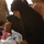 A woman wearing a black hijab/abaya leans over a swaddled infant lying on a white surface, appearing to be in a medical or aid setting. A person in military camouflage uniform is partially visible at the bottom of the frame, suggesting this may be a military-organized medical assistance event. Several other individuals are visible in the background.