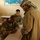 The image shows a military interaction scene inside what appears to be a classroom or administrative room. An Iraqi soldier in desert camouflage is writing on a paper at a desk, while a US Army soldier observes from behind, and a civilian man wearing a keffiyeh stands across the table. A world map with Arabic text is visible on the wall in the background.