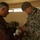 A U.S. Army Special Forces soldier wearing ACU (Army Combat Uniform) and a stethoscope is providing medical assistance or distributing medication to a civilian man. The soldier appears to be examining or handing over items from a box containing what appear to be medical supplies or medications. The setting appears to be an improvised medical aid station in a building, consistent with a humanitarian or civil affairs mission.