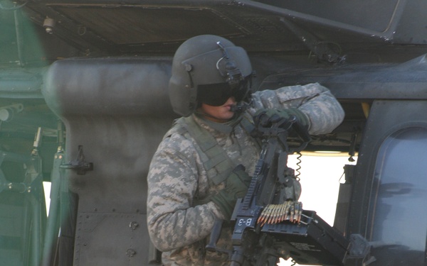 A U.S. Army soldier in Army Combat Uniform (ACU) digital camouflage pattern stands on the landing skid/wheel assembly of a military helicopter, identified by partial text as a Black Hawk. The soldier is wearing a flight helmet with visor and is manning or inspecting a crew-served machine gun mounted on the aircraft door. Ammunition belt is visibly loaded into the weapon system.