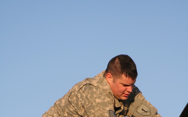A U.S. Army soldier in ACU (Army Combat Uniform) digital camouflage is performing maintenance or inspection work on top of a military vehicle or aircraft. The soldier is leaning forward and working with both hands on mechanical components. The image is taken from a low angle against a clear blue sky, suggesting an outdoor setting during daylight hours.