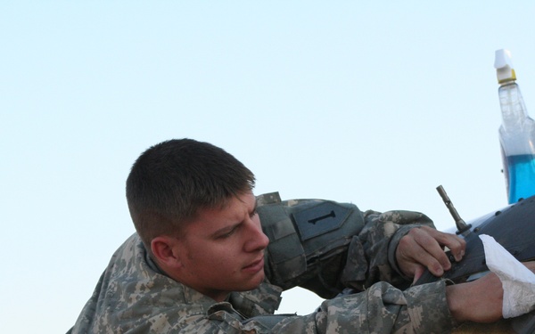 A soldier in US Army ACU (Army Combat Uniform) digital camouflage is leaning over what appears to be a military vehicle windshield, performing maintenance or repair work. He is holding a tool and using a cloth or rag while working on the vehicle's windshield or wiper area. A bottle of blue liquid (likely windshield washer fluid) is visible on top of the vehicle.