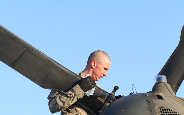 A U.S. Army soldier in Army Combat Uniform (ACU) is performing maintenance or inspection work on top of a military helicopter, likely a UH-60 Black Hawk, focusing on the tail rotor assembly. The soldier is positioned atop the aircraft's tail section, gripping the rotor hub area while wearing tan combat boots and black gloves. The image is taken from a low angle against a clear blue sky.