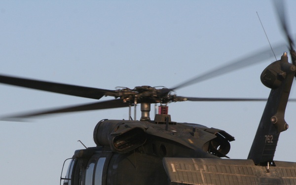 A military helicopter, identifiable as a UH-60 Black Hawk, is captured in flight against a clear, pale blue sky. The aircraft bears the tail number '762' and is painted in a dark olive/black military finish. The main rotor blades show motion blur, indicating active flight at low altitude.