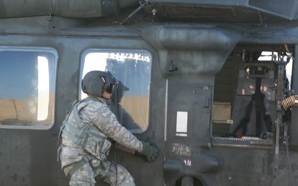 A U.S. Army soldier in full combat gear is boarding or inspecting a military helicopter, identified as a UH-60 Black Hawk, on what appears to be a tarmac or airfield. The soldier is wearing ACU (Army Combat Uniform) pattern camouflage, a flight/combat helmet with communications gear, body armor, and tactical gloves. The helicopter's side door is open, revealing an interior-mounted machine gun with ammunition belt.