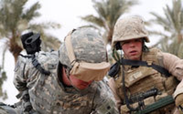 Two military personnel in desert combat gear are crouched over sandy ground, with one soldier closely examining or handling a circular object on the ground that appears to be an IED or landmine with visible wiring. A second soldier in tan/desert MARPAT uniform crouches nearby providing observation. Palm trees are visible in the background, suggesting a Middle Eastern environment.