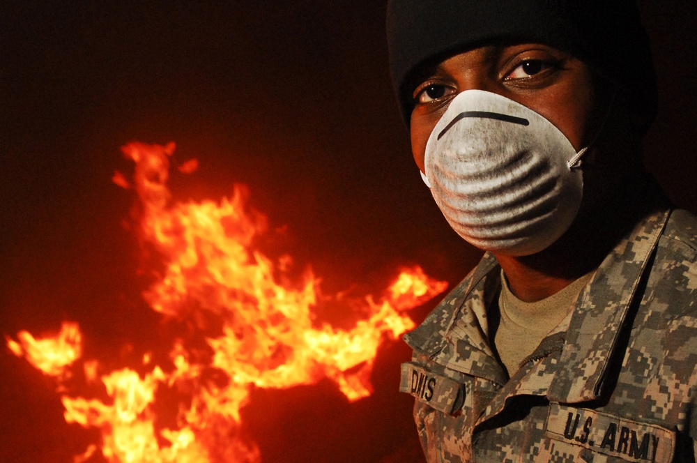 A U.S. Army soldier wearing digital camouflage (ACU pattern) and a white N95-style respirator mask stands in front of a large open fire at night. The soldier is also wearing a dark beanie cap, and a name tape partially reading 'DAVIS' or similar is visible on the uniform. The image appears to document military operations involving burn pits or controlled fire, a practice associated with waste disposal in deployed environments.