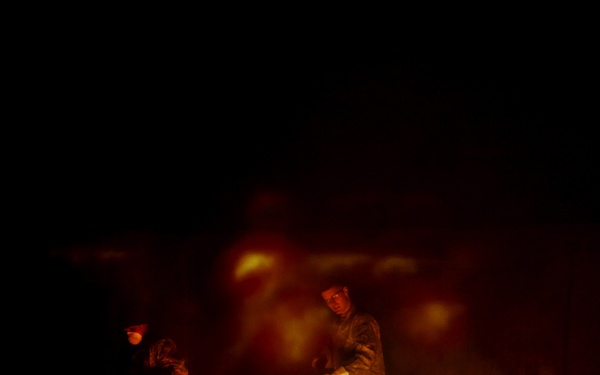 Two individuals wearing military camouflage uniforms are standing near multiple burning containers at night. The scene is illuminated primarily by the flames from what appear to be burn barrels or waste containers. The dark background and fire lighting suggest an outdoor nighttime burn pit operation, commonly associated with military waste disposal.