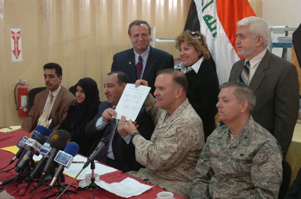 A formal signing or document presentation ceremony is taking place at a table with multiple microphones from various Arabic-language media outlets. The gathering includes civilians in business attire, a woman wearing a hijab, and two individuals in military uniforms — one in desert camouflage and one in U.S. Air Force dress uniform. An Iraqi flag is visible in the background, suggesting this is an official event in or related to Iraq.
