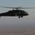 A dark military helicopter is photographed in flight at altitude over an arid, flat terrain. The helicopter displays a red cross medical emblem on its fuselage, identifying it as a medevac or medical evacuation aircraft. The image appears to be taken from another aircraft, with hazy atmospheric conditions visible.