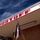 The image shows the exterior overhang of a building labeled 'DUSTOFF' in large red letters on a white fascia, decorated with red cross symbols. A flag hangs limply from a hook beneath the overhang, and the building's exterior wall appears to be plywood or wood paneling. The photo is taken from a low angle looking upward against a blue sky with scattered clouds.
