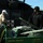 Two individuals are loading or unloading supplies from a John Deere Gator Diesel utility vehicle near a military helicopter marked with a Red Cross symbol, indicating a medical or humanitarian relief operation. One person wears a tan shirt and white cap, while the other is dressed in all black with a dark beanie. The scene appears to take place on a tarmac or open ground in daylight.