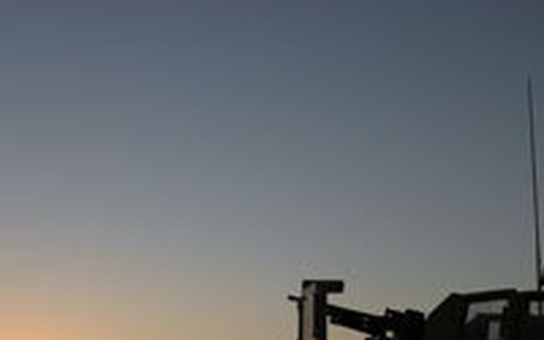 A silhouetted military scene captured at sunset or sunrise, showing armored military vehicles and at least one soldier against a vivid orange and blue sky. The low light conditions render all foreground subjects as dark silhouettes. The flat, open terrain suggests a desert or arid environment.