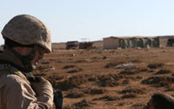 A uniformed military soldier in desert camouflage and helmet is standing face-to-face with a young child in an arid, rural environment. The soldier appears to be engaged in conversation with the child, who is looking up at them. The background shows sparse desert terrain with low structures visible in the distance.
