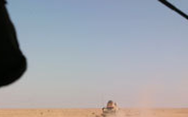 The image is taken from inside a moving vehicle, looking through the windshield across a flat, arid desert landscape. In the middle distance, another vehicle is visible kicking up a dust cloud as it travels across the terrain. A silhouetted figure is visible in the foreground on the left side of the frame.