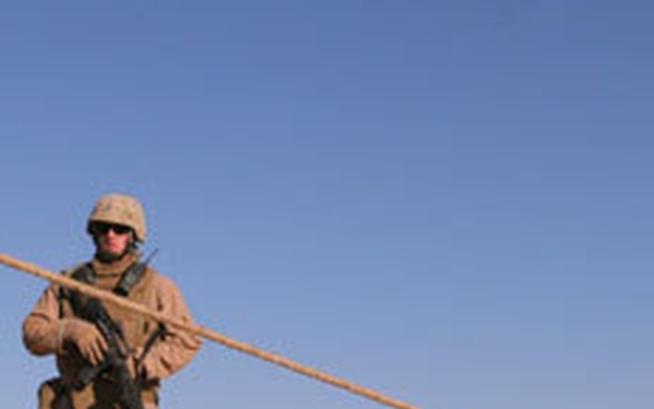 A uniformed soldier stands in an arid desert environment holding a rifle, with ropes or cables extending diagonally across the frame. In the foreground lies a charred or damaged mechanical object on the sandy ground. The scene suggests a post-incident assessment or security perimeter operation in a desert combat zone.