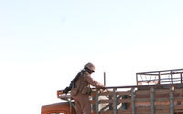An armed soldier in desert camouflage uniform and helmet is climbing onto or inspecting the flatbed of an orange and blue civilian cargo truck in an arid, desert environment. A second individual is visible at ground level near the rear of the truck. The setting appears to be a checkpoint or vehicle inspection in a desert region.