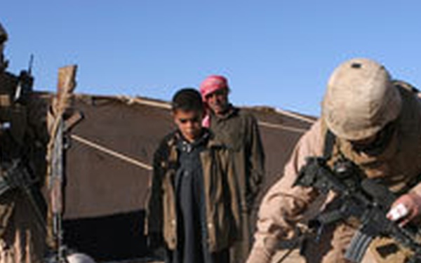 The image shows two armed military personnel in desert camouflage uniforms interacting with a child and an adult civilian in an arid, desert environment. One soldier is crouching down near the child while the other stands guard nearby. A makeshift tent or shelter is visible in the background.