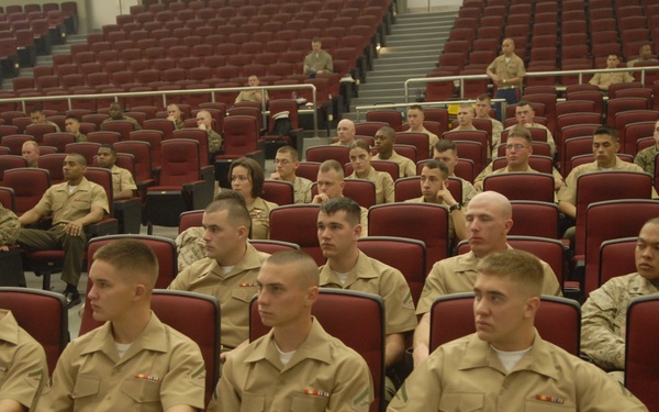 A large group of military personnel, appearing to be U.S. Marines in tan/khaki service uniforms, are seated in a red-seated auditorium or lecture hall. The personnel are arranged in rows facing forward, suggesting a briefing, training session, or formal presentation is taking place or about to begin. The front rows are more densely occupied while the rear of the auditorium has sparser attendance.