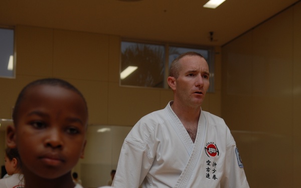 A karate class is in session in a dojo or gymnasium setting. A young child in the foreground is executing a punch technique while a black-belt adult instructor in a white gi observes in the background. Additional students in white uniforms are visible in the background near a mirrored wall.