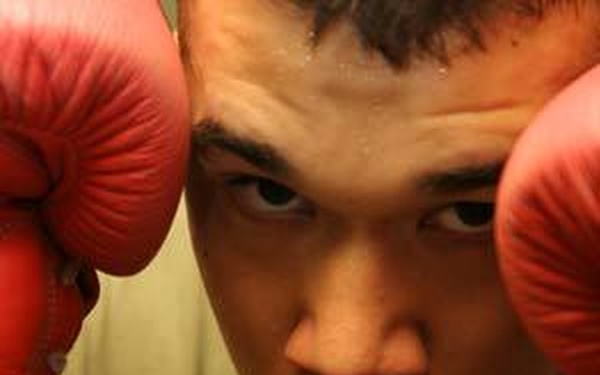 A young male boxer is photographed in a close-up defensive stance with red boxing gloves raised near his face. He is wearing a dark green t-shirt and the gloves appear to be laced, suggesting older-style or traditional boxing equipment. His expression is intense and focused, directed toward the camera.