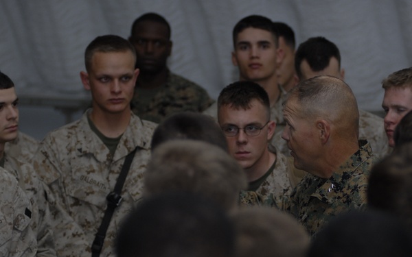 A group of U.S. Marines in desert camouflage (MARPAT) uniforms are gathered in what appears to be a tent or temporary structure. A senior Marine, identifiable by rank insignia and a shaved head, appears to be addressing or briefing the assembled junior Marines. The scene suggests a formal or semi-formal military briefing or address.