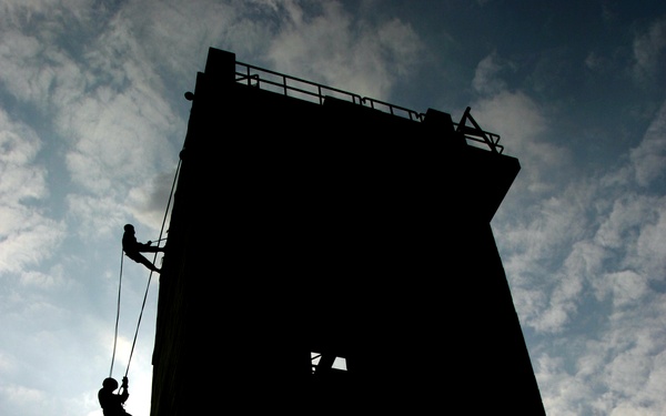 A dramatic silhouette photograph showing two individuals rappelling down the face of a tall rectangular training tower. The image is backlit against a partly cloudy sky, creating strong contrast between the dark structure and the bright background. A chain-link fence is visible in the foreground, suggesting this is a controlled training facility.