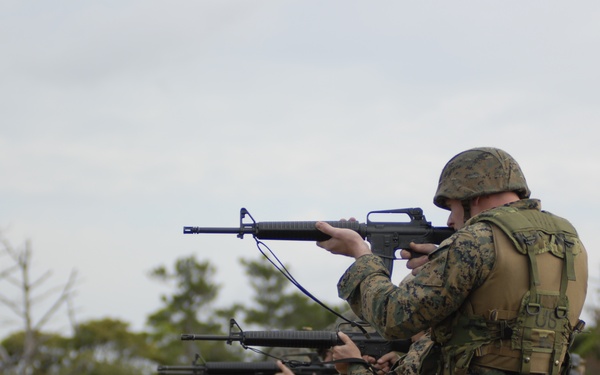 A line of military personnel in MARPAT (Marine pattern) camouflage uniforms and tactical gear are conducting a rifle firing exercise at an outdoor range. The soldiers are in a standing shooting stance, aiming M16-style rifles downrange. At least one observer or instructor is visible in the background holding papers.