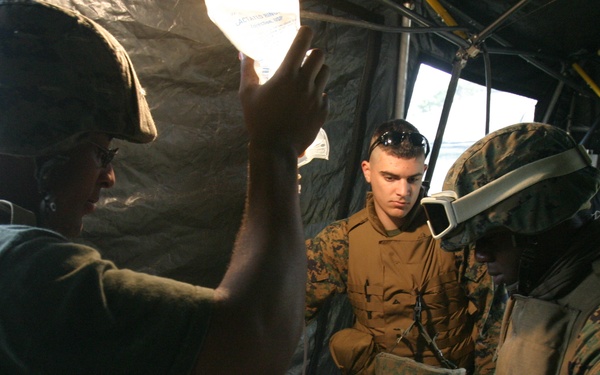 Military medical personnel in camouflage uniforms are performing a field medical procedure or training exercise on a casualty lying on a stretcher inside a tactical tent or shelter. The patient has visible simulated wound makeup on the torso with medical leads/wires attached. Three personnel are actively engaged around the patient, with one using what appears to be a handheld medical device.