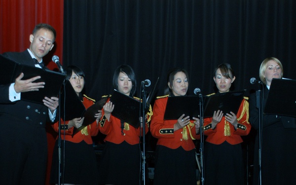 A formal musical performance on a stage featuring a male emcee or soloist in a tuxedo and four female singers wearing matching red military-style jackets with gold trim over black skirts and black knee-high boots. A fifth woman in black attire stands at the far right, and additional musicians in red uniforms are visible in the background. The group is performing in front of black curtains with a red curtain visible on the left side.