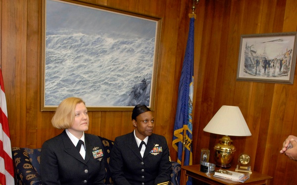 Four U.S. Navy personnel are gathered in what appears to be a formal office or meeting room with wood-paneled walls. Three individuals in dark Navy dress uniforms are seated on a sofa, while a senior officer in tan/khaki service uniform stands and appears to be speaking to the group. The setting suggests an official meeting or briefing in a flag officer's office.