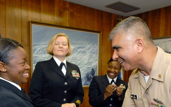 A formal military ceremony or recognition event taking place in a wood-paneled office setting. A male Navy enlisted sailor in khaki uniform is shaking hands with a female sailor in dress blues, while two other female Navy officers look on smiling. The atmosphere is celebratory, suggesting an award, promotion, or commendation event.