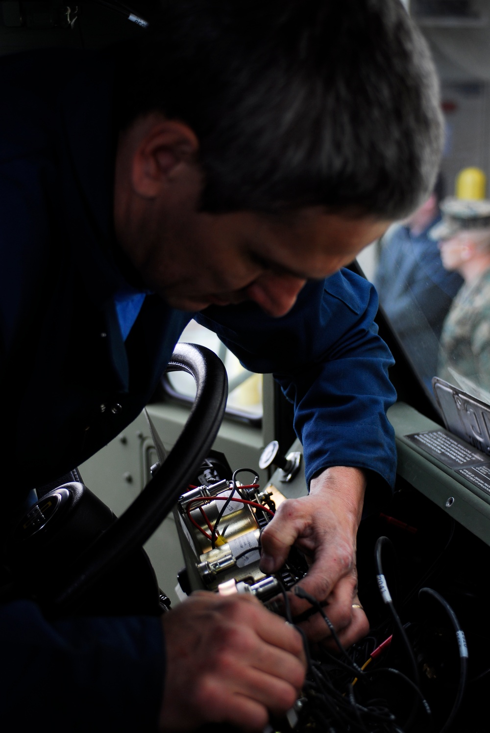 A man in a blue uniform is working on electrical or mechanical components inside what appears to be a vehicle or equipment cab. He is leaning over a console area, manipulating wiring and small components with both hands. The background shows additional personnel and equipment panels.