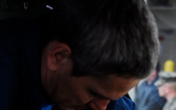 A man in a blue uniform is working on electrical or mechanical components inside what appears to be a vehicle or equipment cab. He is leaning over a console area, manipulating wiring and small components with both hands. The background shows additional personnel and equipment panels.