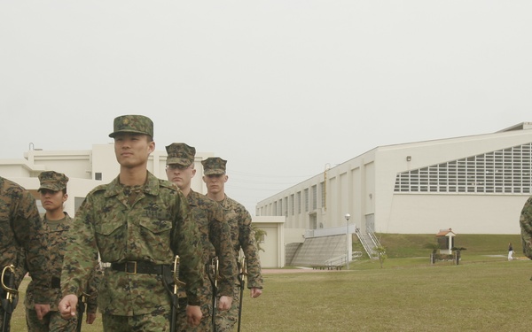 A group of military personnel in camouflage uniforms are marching across a grass field, carrying ceremonial swords or sabers at their sides. The scene appears to depict a joint military drill or ceremonial exercise, likely involving both U.S. Marines and Japanese Ground Self-Defense Force members based on the distinct uniform patterns visible. The setting appears to be a military installation with white institutional buildings in the background.