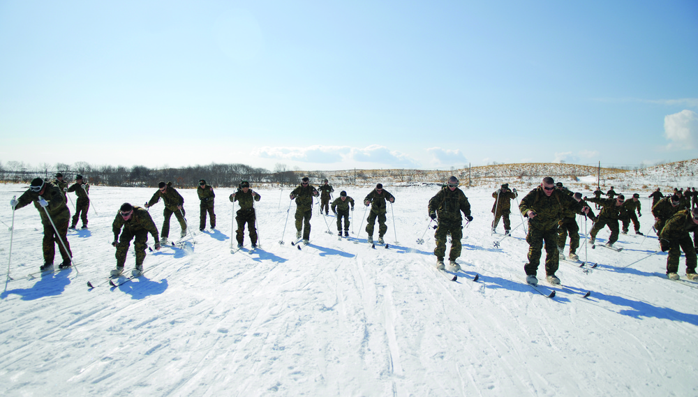 A large group of military personnel in camouflage uniforms are conducting cross-country skiing training on a snow-covered flat terrain. The soldiers are equipped with ski poles and appear to be in various stages of a skiing drill or exercise. The setting appears to be a cold-weather military training environment with sparse trees and rolling hills visible in the background.