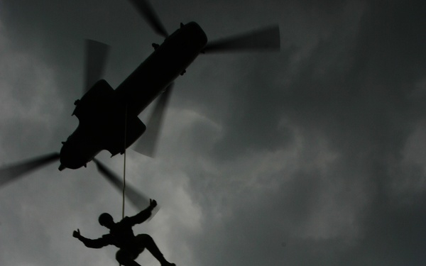 A dramatic silhouette photograph taken from below showing multiple personnel fast-roping or rappelling from a helicopter against an overcast, stormy sky. At least three individuals are visible suspended on a single rope descending from the aircraft. The low-angle, backlit composition renders all subjects as dark silhouettes against the grey cloud-filled background.