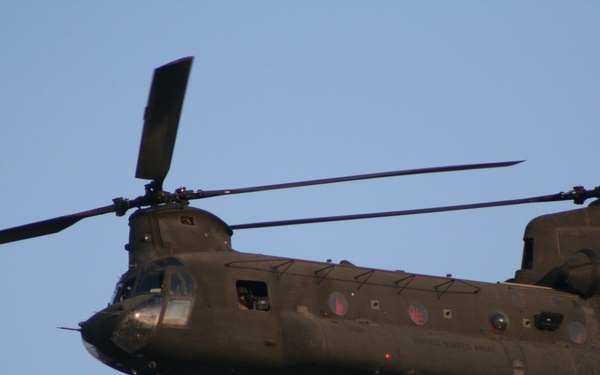 A United States Army CH-47 Chinook tandem-rotor helicopter is photographed in flight against a clear blue-grey sky. The aircraft is olive drab in color and appears to be in level flight, viewed from a slightly below-and-side angle. Text on the fuselage reads 'United States Army'.