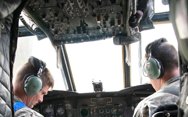 Two military personnel in US Army ACU (Army Combat Uniform) camouflage are seated in the cockpit of a large military helicopter, identifiable by the 'VERTOL' placard visible on the center console, indicating a Boeing CH-47 Chinook or similar Vertol-manufactured aircraft. Both crew members are wearing green aviation headsets and are engaged in cockpit operations, with one reaching toward the center console controls. The instrument panel shows multiple analog gauges, green indicator lights, and digital displays consistent with a military rotary-wing aircraft.