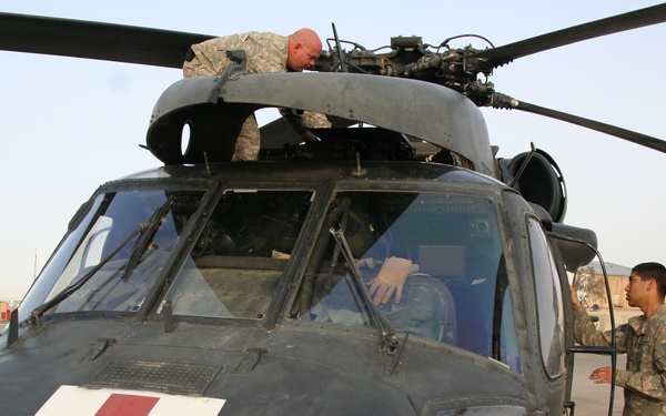 A military medevac helicopter, identified by the prominent Red Cross marking and number '888' on its nose, is undergoing maintenance or pre-flight inspection on a tarmac. Three soldiers in U.S. Army combat uniforms are working on the aircraft — one on top of the fuselage, one inside the cockpit, and one standing alongside the helicopter. The setting appears to be a forward operating base or military airfield, likely in a Middle Eastern environment based on the terrain and infrastructure visible in the background.