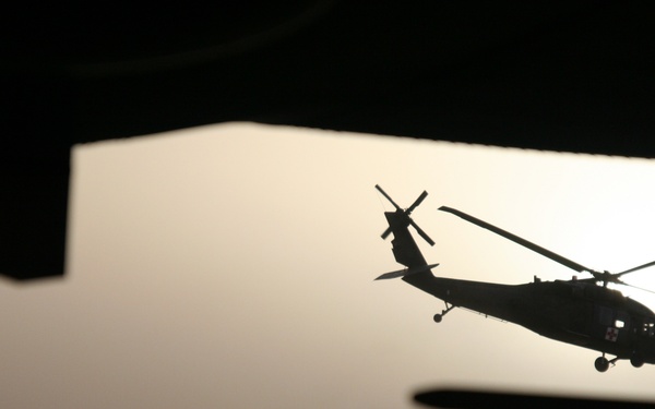 A military helicopter, consistent with a UH-60 Black Hawk configuration, is photographed in silhouette against a hazy, overcast sky. The image is taken from inside a vehicle, with structural framing elements visible at the top and bottom of the frame. A red cross marking is faintly visible on the helicopter's fuselage, suggesting a medevac or medical evacuation role.