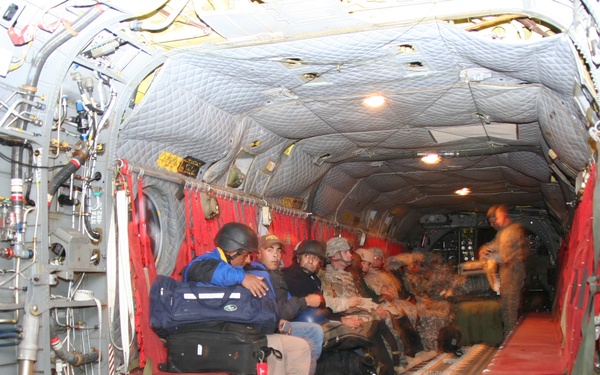 The image shows the interior of a military cargo aircraft, likely a CH-47 Chinook or similar transport helicopter, with passengers seated along the side walls on red netting seats. A mix of civilians and military personnel in camouflage uniforms are seated with luggage and gear. The cargo bay is illuminated by overhead lights and shows typical military aircraft interior features including exposed hydraulic lines, cargo floor tracks, and quilted insulation panels.