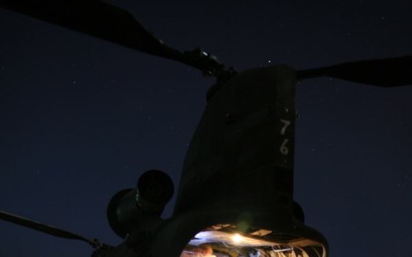 A large tandem-rotor military helicopter, identifiable as a CH-47 Chinook, is photographed at night with its rear cargo ramp lowered and interior lights illuminated. Personnel in military uniforms are visible inside the aircraft and on the ramp, suggesting a loading, unloading, or staging operation. The number '76' is visible on the rear fuselage, and concrete barrier walls (T-walls/HESCO) are visible in the background, indicating a forward operating base or military installation.